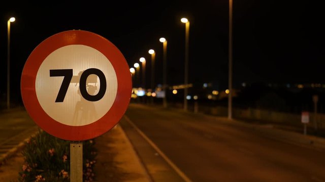 At night on the Spanish national road N332. A sign shows a speed limit of 70. Vehicles drive past, blurred in the background with beautiful bokeh.