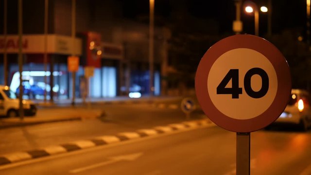 In the city of the Spanish city of Torrevieja at night lit by lanterns. A sign shows a speed limit of 40 in front of a roundabout. Vehicles drive through the circle. Background Bokeh.