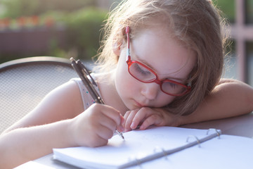 girl with glasses writes in a notebook. sunny soft background. doing homework outdoors