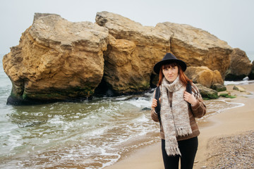 Portrait of a young red-haired woman in a hat and a scarf with a backpack against the background of the rocks against the beautiful sea, on the coast, on the horizon. Tourism, rest, lifestyle.