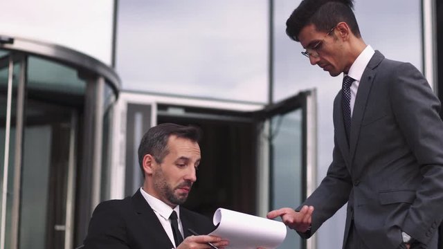 Male adult colleagues in suits talking outdoors, one is on wheelchair, business communication.