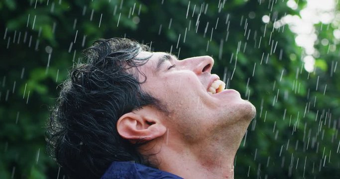 Slow motion of young carefree handsome young man wearing protection cape is feeling free under the rain on a background of green trees.