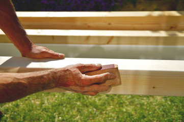 man's hand smoothing natural wooden beam using sandpaper while he is  working outside. Sanding of...