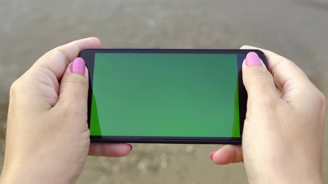 Closeup top view of empty blank green screen of mobile phone in hands of woman standing at summer beach. Real time 4k video footage.