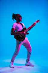 Fototapeta premium Young african-american musician playing the guitar like a rockstar on blue studio background in neon light. Concept of music, hobby. Joyful attractive guy improvising. Retro colorful portrait.