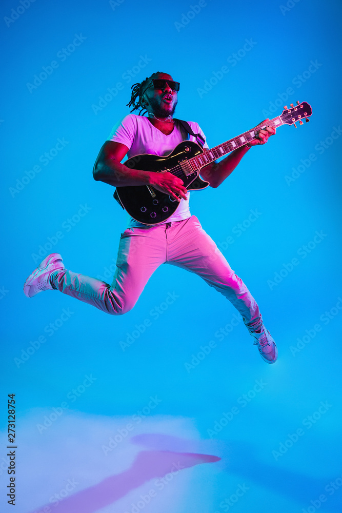 Wall mural Young african-american musician playing the guitar in jump like a rockstar on blue studio background in neon light. Concept of music, hobby. Joyful attractive guy improvising. Retro colorful portrait.