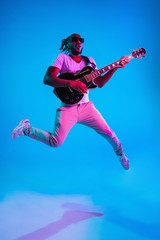 Young african-american musician playing the guitar in jump like a rockstar on blue studio...