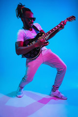 Fototapeta premium Young african-american musician playing the guitar like a rockstar on blue studio background in neon light. Concept of music, hobby. Joyful attractive guy improvising. Retro colorful portrait.