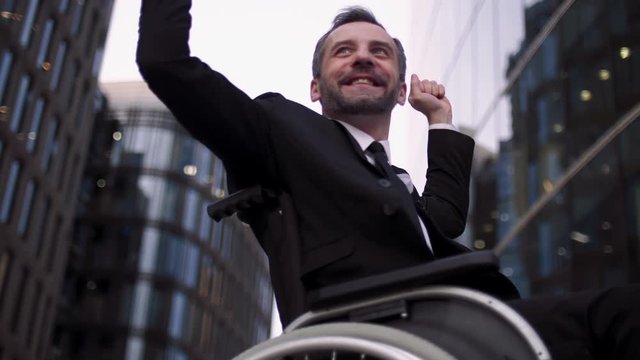 Disabled attractive invalid buisnessman on wheelchair outdoor. Handicapped man near glass building offices. Happy emotion portrait, hands up, win.