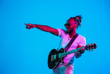 Obraz premium Young african-american musician playing the guitar like a rockstar on blue studio background in neon light. Concept of music, hobby. Joyful attractive guy improvising. Retro colorful portrait.