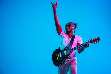 Young african-american musician playing the guitar like a rockstar on blue studio background in...
