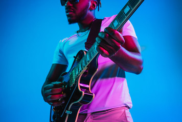 Young african-american musician playing the guitar like a rockstar on blue studio background in neon light. Concept of music, hobby. Joyful attractive guy improvising. Retro colorful portrait. - obrazy, fototapety, plakaty