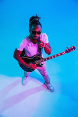 Fototapeta na wymiar Young african-american musician playing the guitar like a rockstar on blue studio background in neon light. Concept of music, hobby. Joyful attractive guy improvising. Retro colorful portrait.