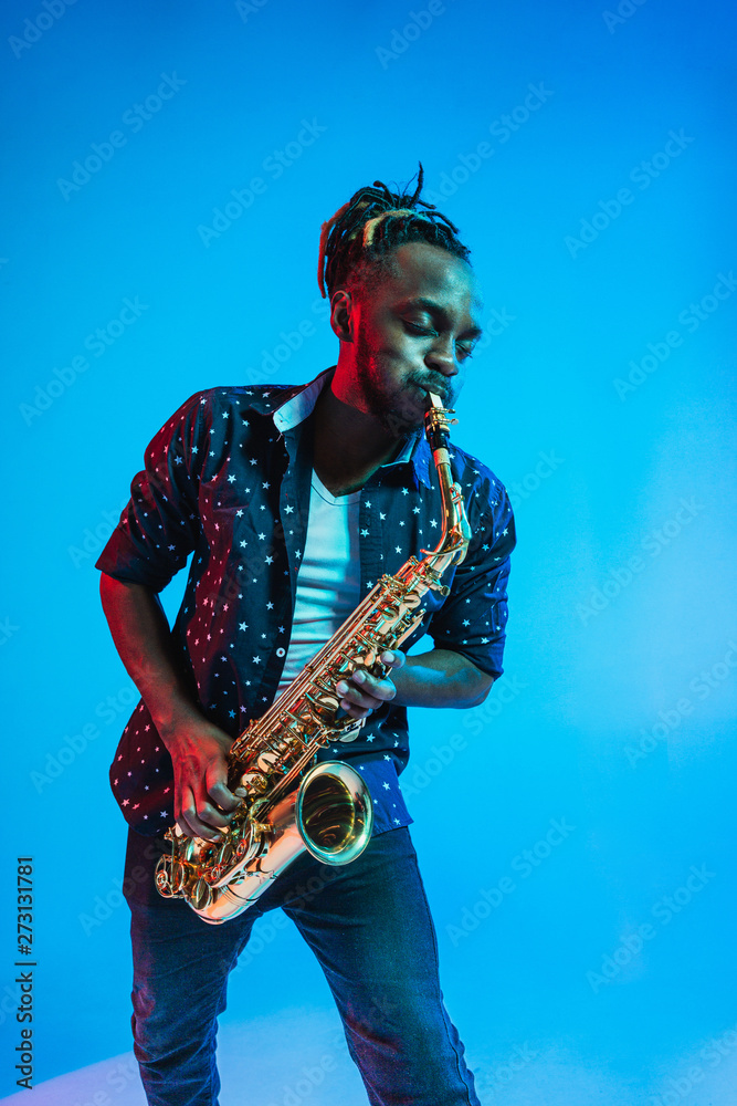 Wall mural Young african-american jazz musician playing the saxophone on blue studio background in trendy neon light. Concept of music, hobby. Joyful attractive guy improvising. Retro colorful portrait of artist