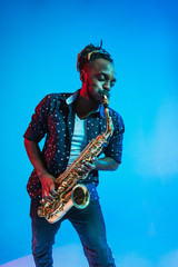 Fototapeta na wymiar Young african-american jazz musician playing the saxophone on blue studio background in trendy neon light. Concept of music, hobby. Joyful attractive guy improvising. Retro colorful portrait of artist