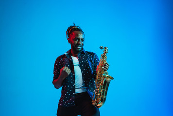 Young african-american jazz musician playing the saxophone on blue studio background in trendy neon...