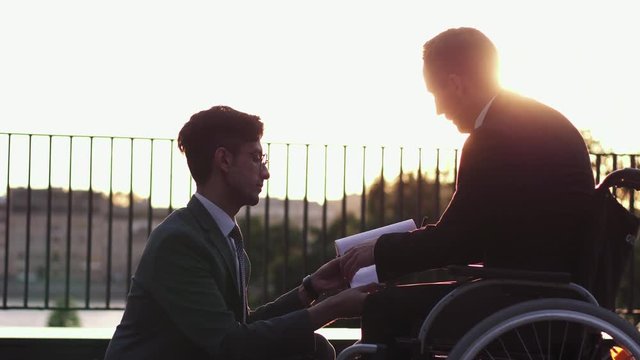 Male adult colleagues in suits talking outdoors, one is sitting on the wheelchair, business communication, front of sunset.