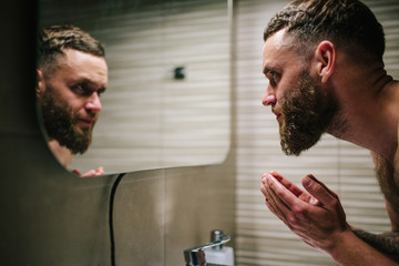 Young bearded hipster man washing his face with water in the bathroom while looking himself in the mirror.
