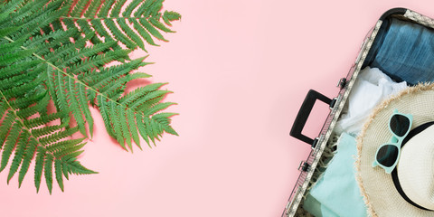 Leaf of fern, tropical detox water and open suitcase with clothes on pastel pink background. Top view with copy space. Summer concept travel.