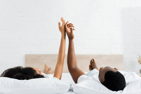 happy african american man touching hands with girlfriend, lying on white bed in bedroom