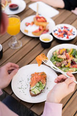 Сompany friends met at lifestyle breakfast in cafe. On the wooden table are plates with food. Salmon on bread with herbs. Black coffee and orange juice. Veggie raw salad. Granola with fruit. Poached.