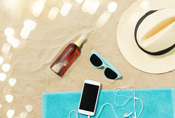 vacation and summer holidays concept - smartphone with earphones on towel, straw hat, sunglasses, flip flops and bottle of sunscreen oil on beach sand