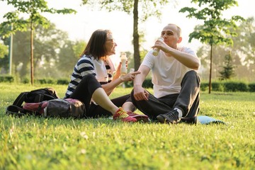 Happy mature couple sitting in the park on fitness mat, resting drinking yogurt after sports exercises.