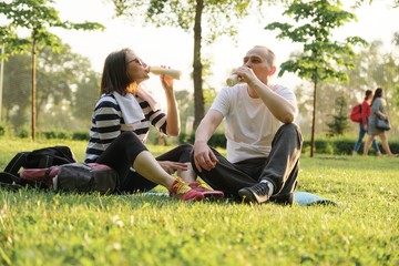 Happy mature couple sitting in the park on fitness mat, resting drinking yogurt after sports exercises.