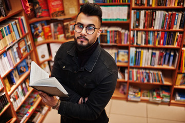 Tall smart arab student man, wear on black jeans jacket and eyeglasses, at library with book at hands.