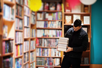 Tall smart arab student man, wear on black jeans jacket and eyeglasses, at library with stack of books.