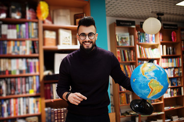 Tall smart arab student man, wear on violet turtleneck and eyeglasses, at library holding earth...