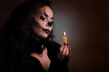 Obraz premium Makeup for Halloween. Portrait of a sexy brunette girl in black clothes with a scary art on her face. Female hands with a burning candle. Pensive look on the flame. Concept in the devilish style.