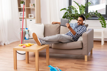 people, housework and housekeeping concept - indian man in headphones listening to music on tablet computer after home cleaning