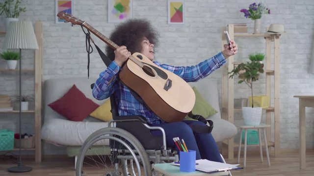 disabled african american woman with an afro hairstyle in a wheelchair takes a selfie with an acoustic guitar