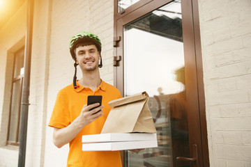 Young man in yellow shirt delivering pizza using gadgets to track order at the city's street. Courier using online app for receiving payment and tracking shipping address. Modern technologies.