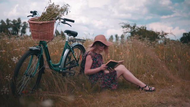 Beautiful Woman Reading Book.Woman In Dress Relaxing.Blonde Woman In Sunglasses.Woman Sitting On Green Grass Field And Reading Book.Beautiful Girl In Hat.Girl With Bicycle.Travel Girl Sitting On Grass
