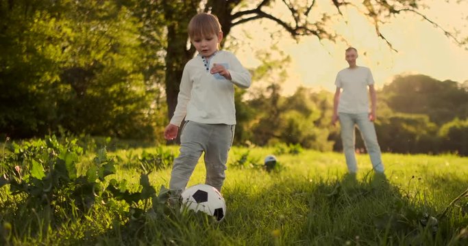 Father And Child Play Football Standing On The Field At Sunset. The Boy Strikes The Goal. The Father Of The Goalkeeper Is On The Gate, The Child Strikes The Ball