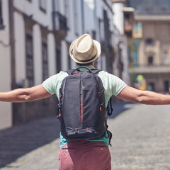  European man in a sun hat is travelling along the narrow street of a European town. His arms are open wide.