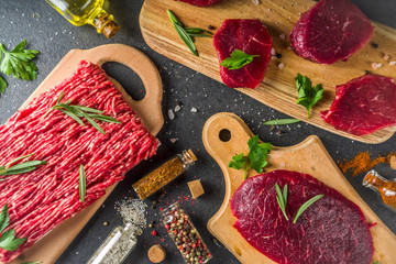 Different raw steak and minced beef meat with herbs and spices for cooking, black concrete or stone...