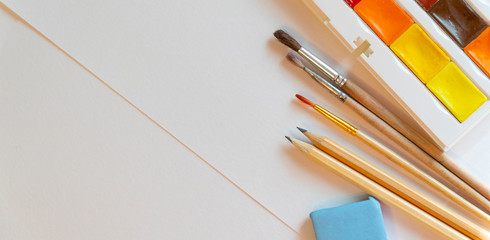 pencils, brushes and watercolor paints on white paper background top view