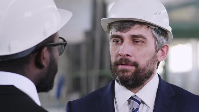 Tilt up shot of Caucasian factory director staying opposite African American workmate and discussing plan of work