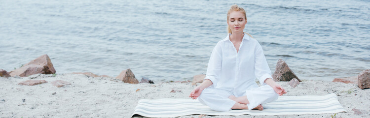 Fototapeta na wymiar panoramic shot of peaceful blonde young woman with closed eyes practicing yoga near river