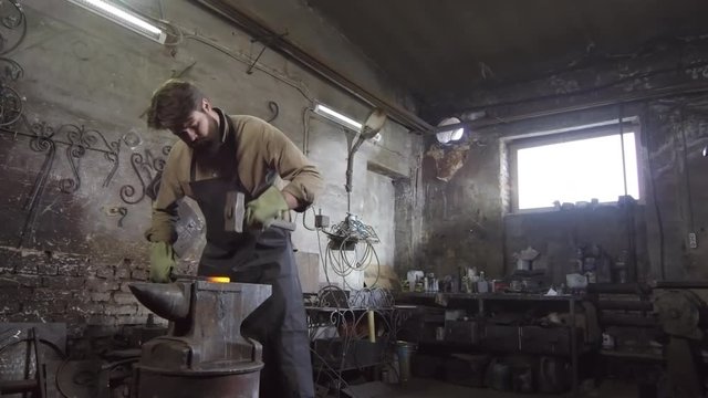 Tracking left of bearded blacksmith wearing apron and gloves standing at anvil and forging hot iron billet with big hammer
