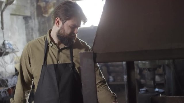 Caucasian bearded blacksmith in safety apron and gloves standing at stove and checking molting metal billets lying in burning coals