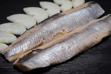 Salted boneless fillets of wild-caught Atlantic Herring and sliced Yellow Onion arranged on natural black stone background. Clupea harengus.