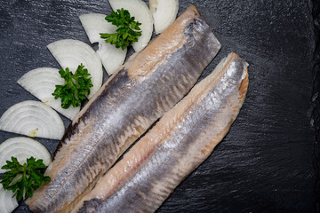 Delicious Salted Herring garnished with Yellow Onion slices and Fresh Parsley. Natural black stone background. Clupea harengus.