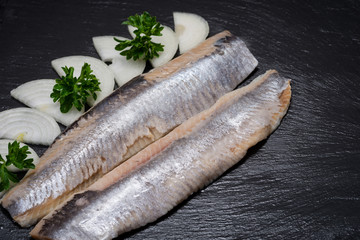 Delicious Salted Herring garnished with Yellow Onion slices and Fresh Parsley. Natural black stone background. Clupea harengus.