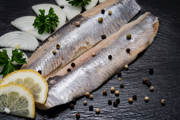 Salted Deboned Herring Fillets garnished with Yellow Onion, Lemon, Fresh Parsley and Peppercorns. Natural black stone background. Clupea harengus.