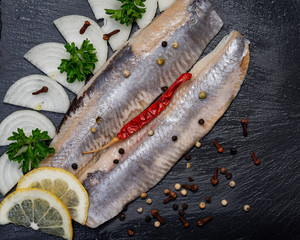 Salted Herring Fillets garnished with Yellow Onion, Lemon, Fresh Parsley and Dried Chili Pepper. Natural black stone background. Clupea harengus.