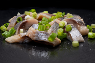 Slices of Salted Boneless wild-caught Atlantic Herring garnished with Chopped Green Onion Scallions. Black natural stone background. Clupea harengus. 